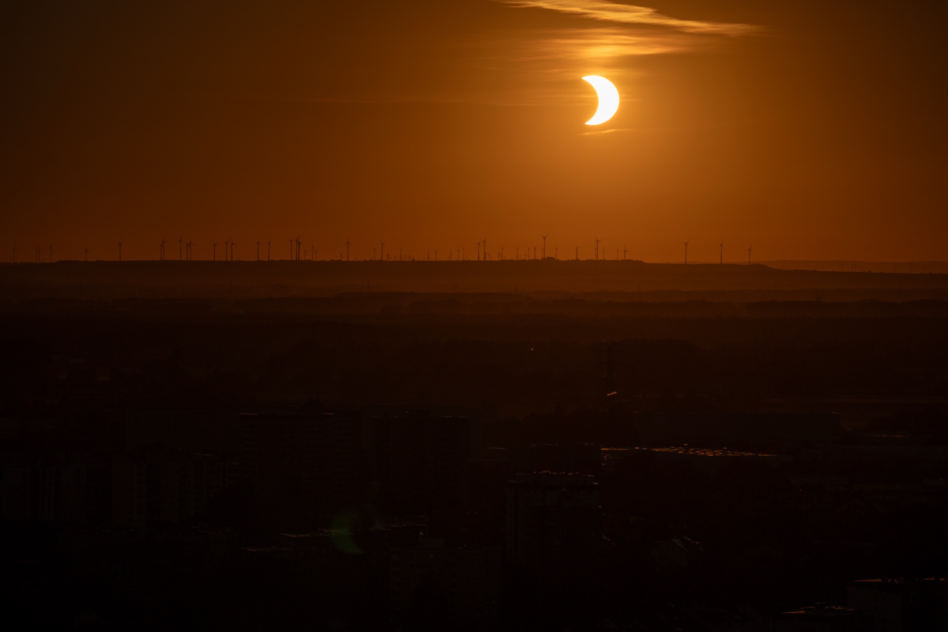 Partielle Sonnenfinsternis als leuchtende Sichel über der Silhouette der Windräder am Horizont, tief oranger Himmel.